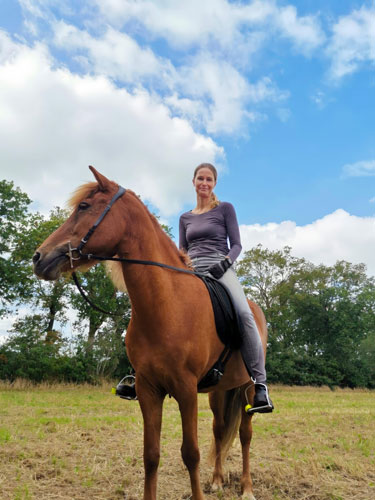 Eine Frau sitzt auf einem braunen Pferd auf einer offenen Wiese, umgeben von Bäumen und unter einem teilweise bewölkten blauen Himmel