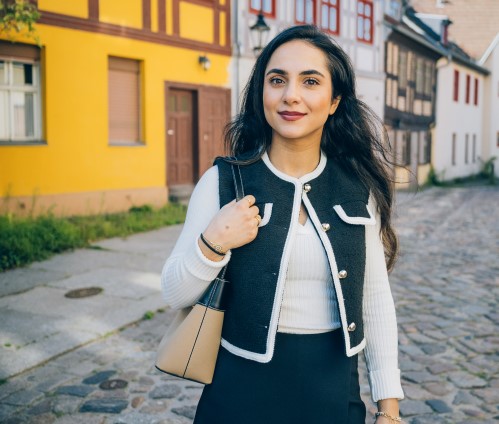 Eine Frau mit langen dunklen Haaren steht auf einer Kopfsteinpflasterstraße in einer Altstadt mit Fachwerkhäusern und trägt eine weiße Strickjacke mit dunkler Weste darüber sowie eine helle Handtasche.