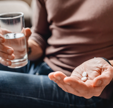 Nahaufnahme einer Person, die in einer Hand ein Wasserglas und in der anderen Hand zwei weiße Pillen hält.